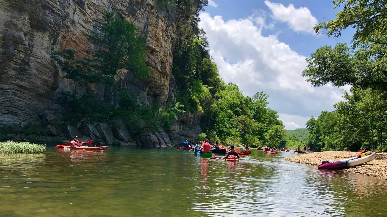 over 10 people in canoes in the river besides a cliff